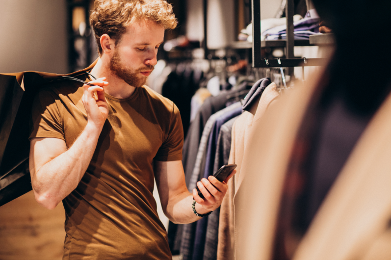 Homme faisant du shopping dans un magasin de vêtements, et recherche des informations sur un article sur le net via son smartphone